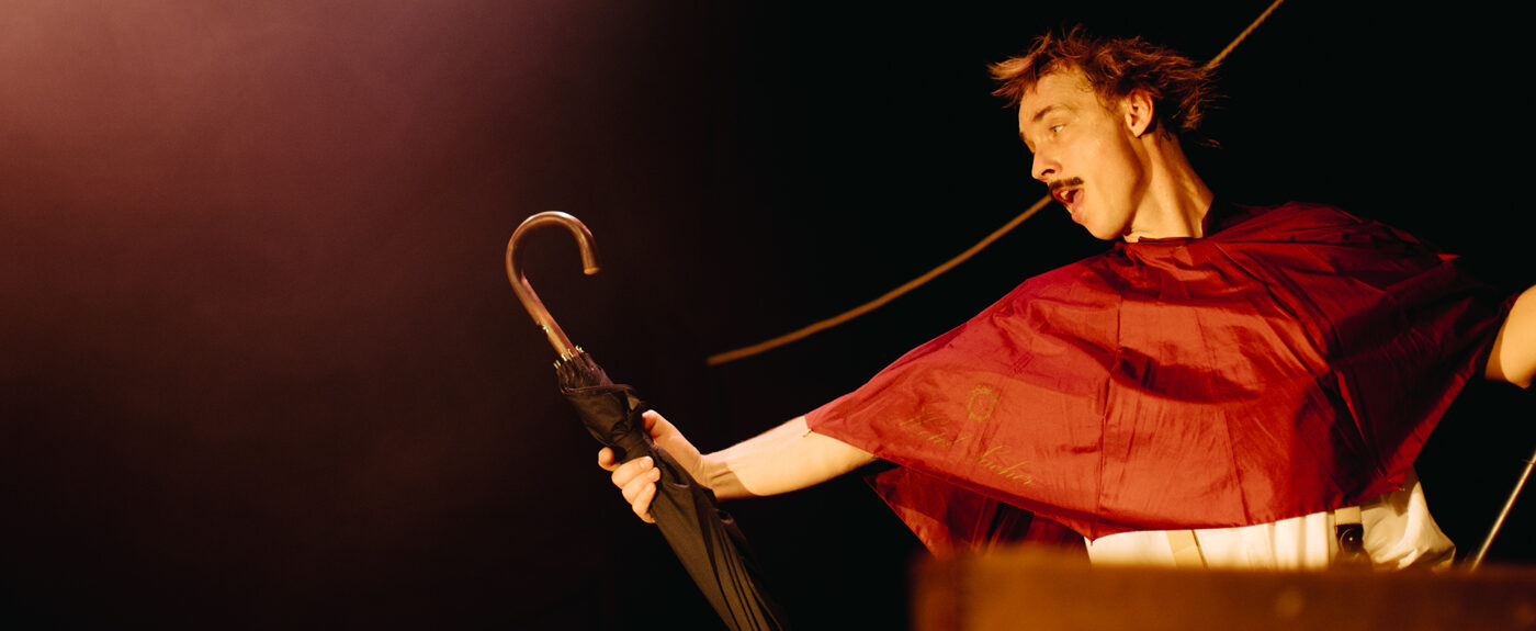 Ein Mann mit Schnurrbart und rotem Umhang hält einen schwarzen Regenschirm auf einer Bühne.