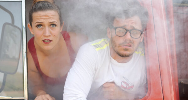 Eine Frau und ein Mann blicken aus dem Fenster eines roten Fahrzeugs, umgeben von Rauch.